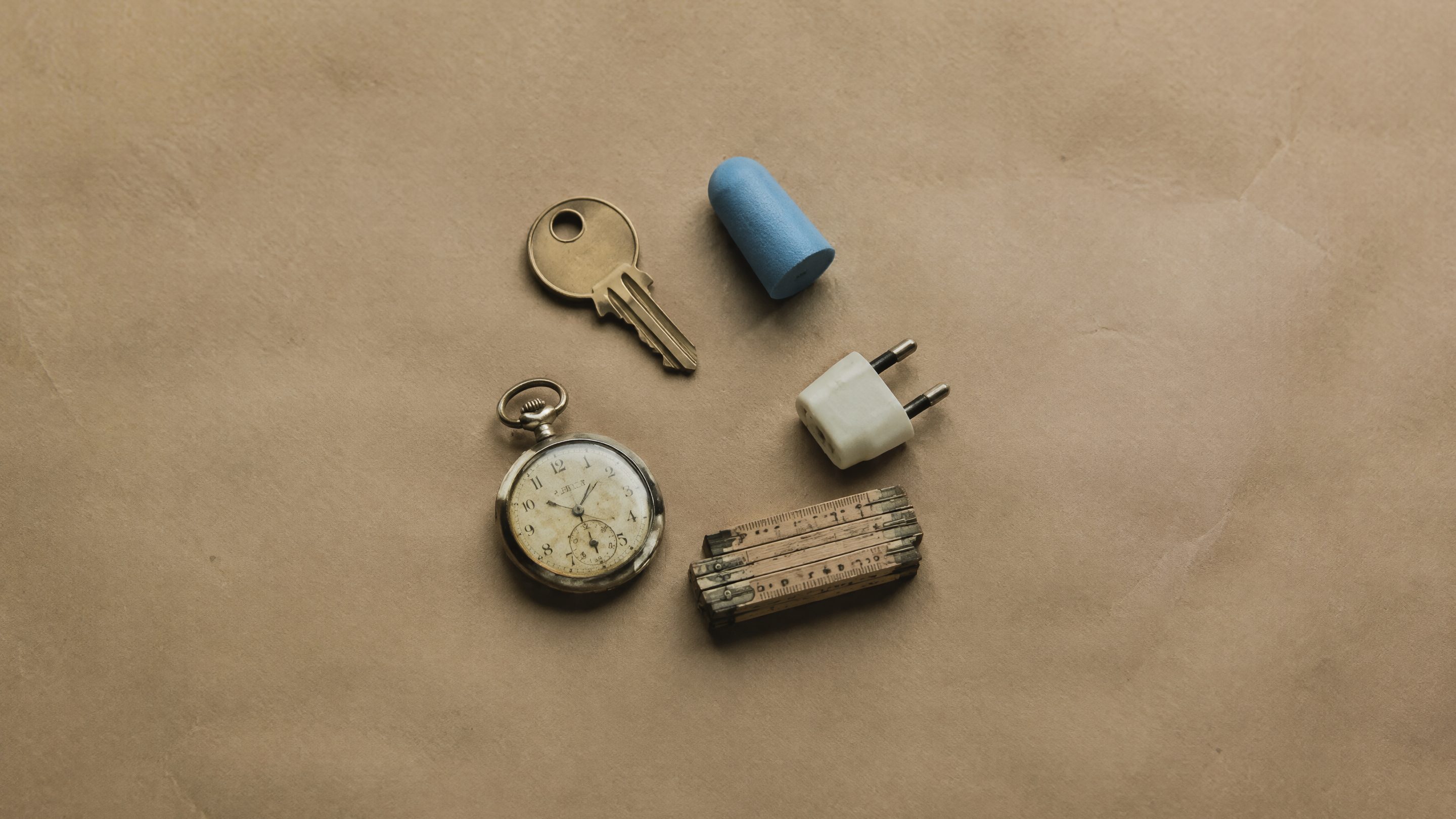 Cinq petits objets disposés en cercle sur du papier kraft : une clé en laiton, un bouchon d'oreille, une fiche électrique, un mètre pliant et une montre de poche usée.