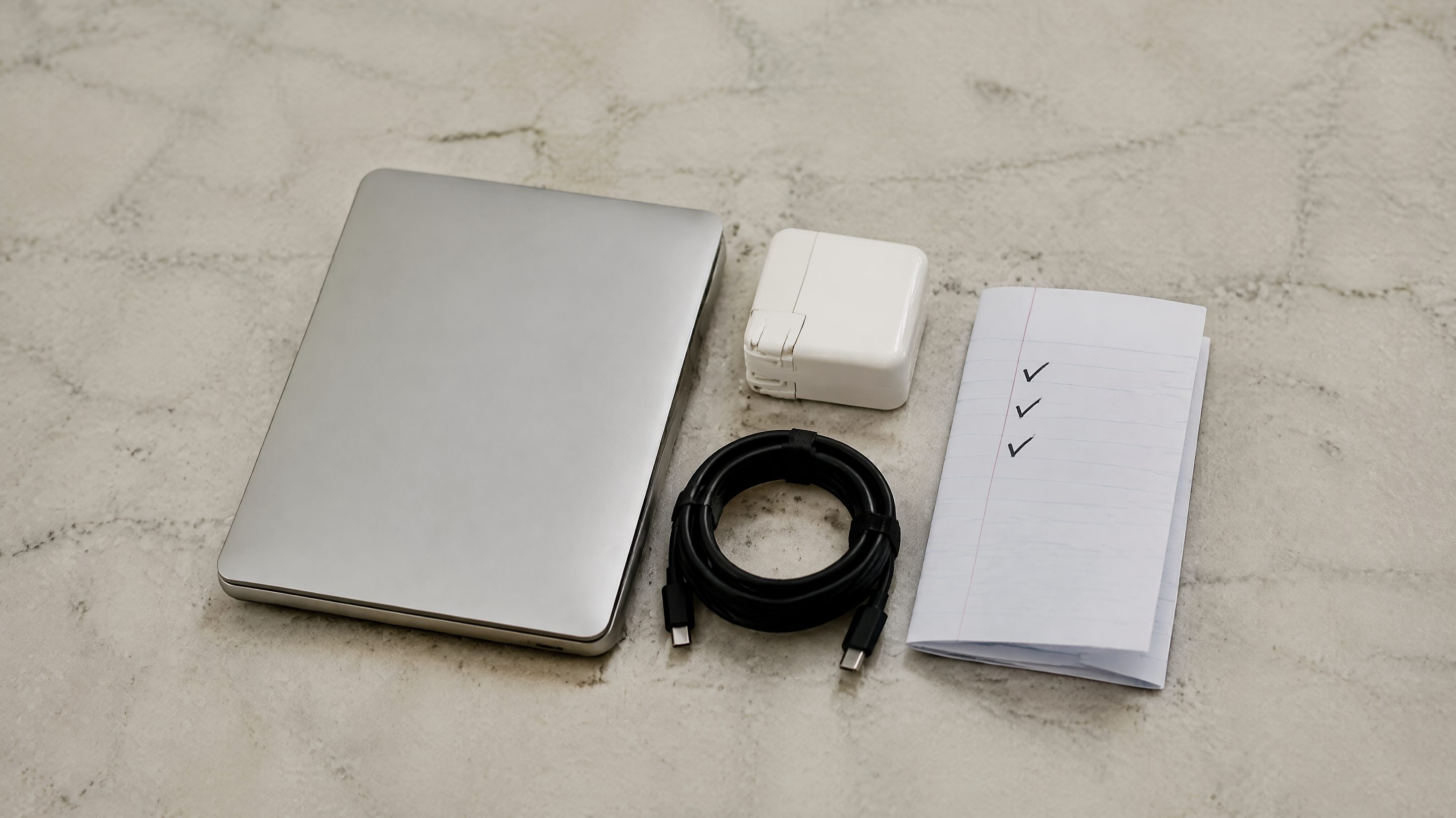 Sur du marbre gris clair, le coin d'un ordinateur portable fermé, un petit chargeur blanc, un câble noir enroulé et une liste manuscrite cochée trois fois.