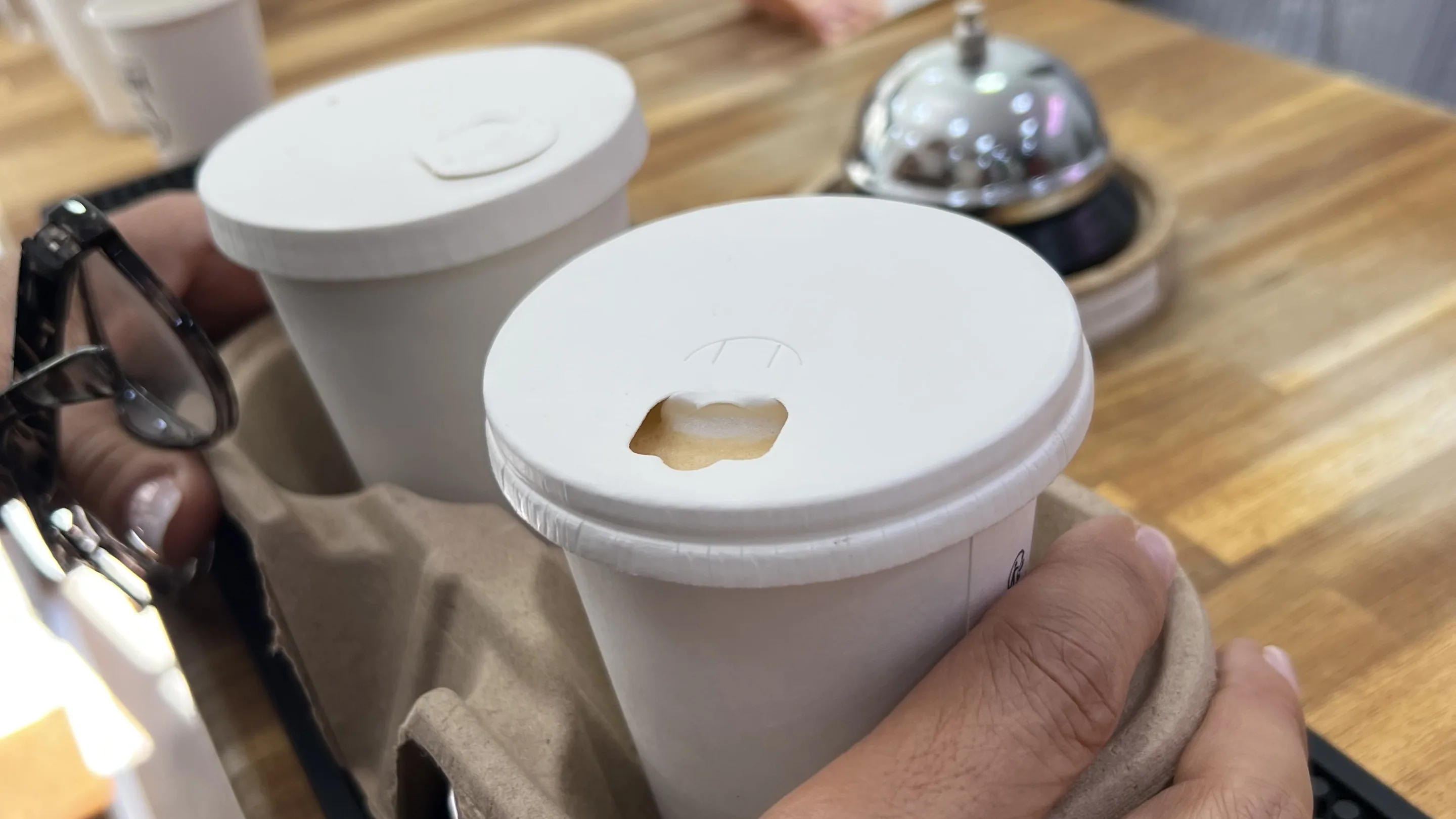 Deux mains tiennent un porte-gobelets en carton avec deux gobelets blancs a couvercle, dont un laisse deborder un peu de cafe, au-dessus d'un comptoir en bois ou sont poses une sonnette metallique et des gobelets empiles