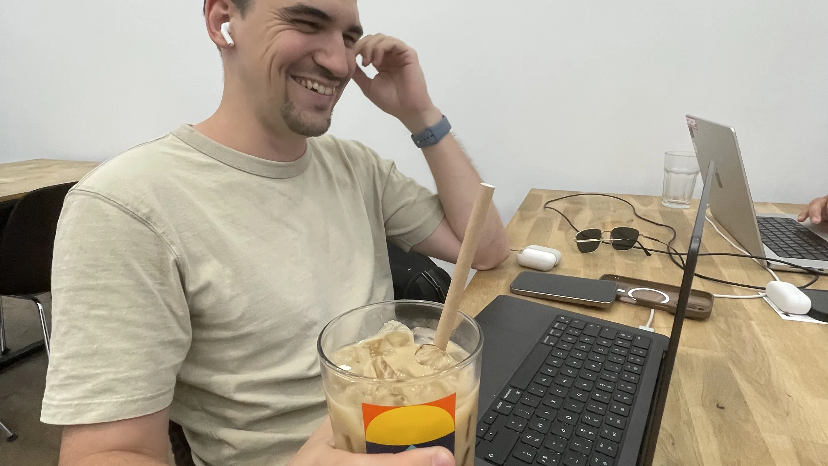 Un homme souriant, assis à une grande table en bois du café, tient à deux mains un verre de latte glacé avec une paille. Son ordinateur portable est ouvert à côté de lui, un deuxième ordinateur et des lunettes de soleil occupent le reste de la table, et une affiche colorée est accrochée au mur clair derrière lui.