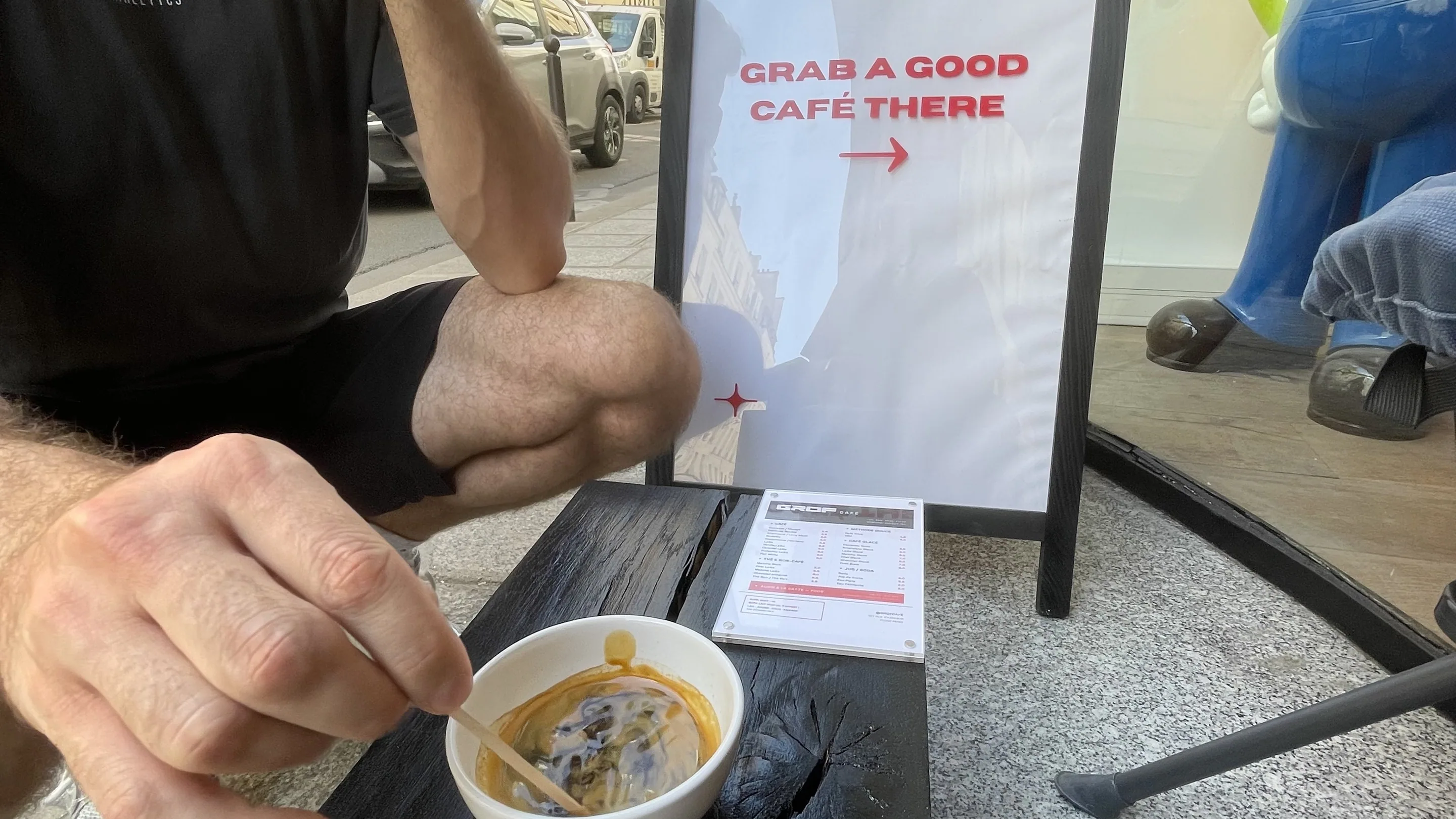Sur le trottoir devant GROF CAFÉ, un homme en t-shirt noir, assis, remue un café servi dans un bol blanc posé sur un billot de bois brûlé. À côté du bol, la carte du café. Derrière, un chevalet blanc où on lit « GRAB A GOOD CAFÉ THERE » avec une flèche rouge, une vitrine, et la rue parisienne avec ses immeubles et ses voitures garées.