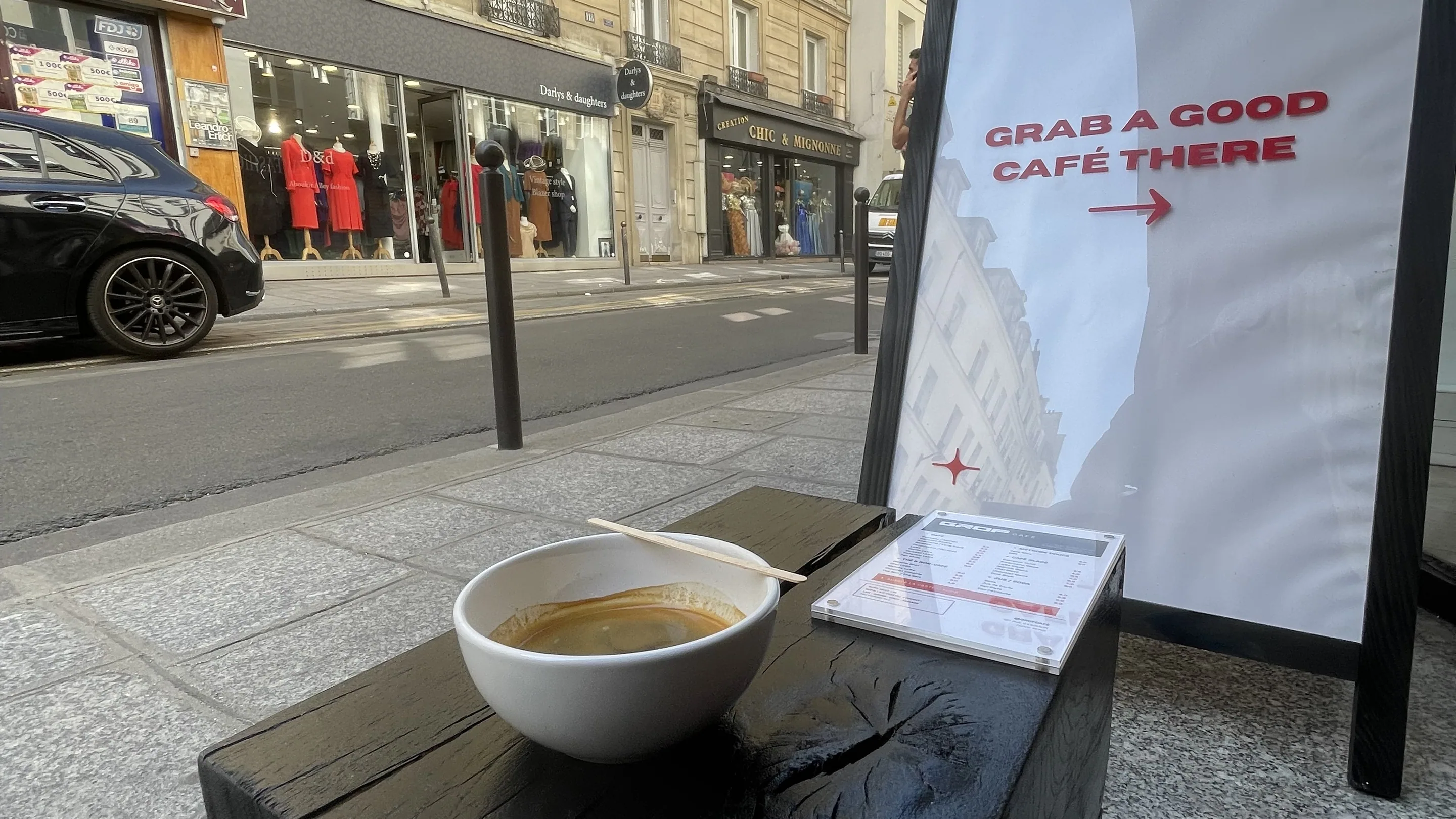 Sur le trottoir du 127 rue d'Aboukir, un bol de café posé sur un bloc de bois noirci qui sert de table, avec la carte du café à côté. Un chevalet porte la mention « Grab a good café there » et une flèche vers l'entrée. En face, la rue ensoleillée, ses immeubles haussmanniens, un tabac et des boutiques de prêt-à-porter.