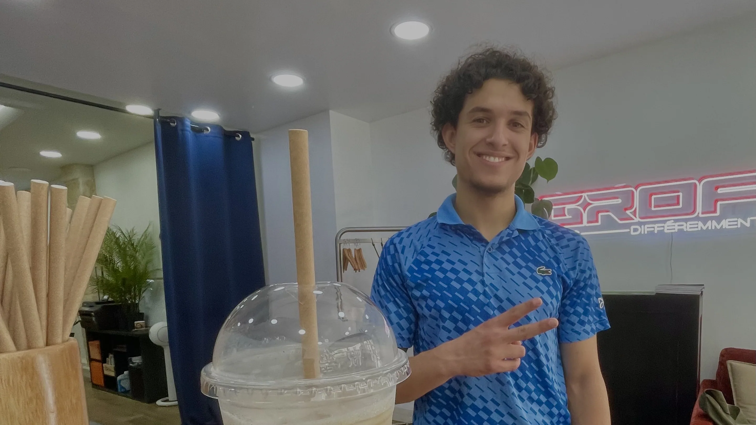 Une personne aux cheveux bouclés courts, en polo bleu, sourit et lève deux doigts derrière le comptoir de GROF CAFÉ. Au premier plan, un grand gobelet transparent d'iced latte, couvercle bombé et paille en papier, posé sur un tapis de bar noir. Au fond, l'enseigne lumineuse GROF « DIFFÉREMMENT », une plante verte et un fauteuil rouge.