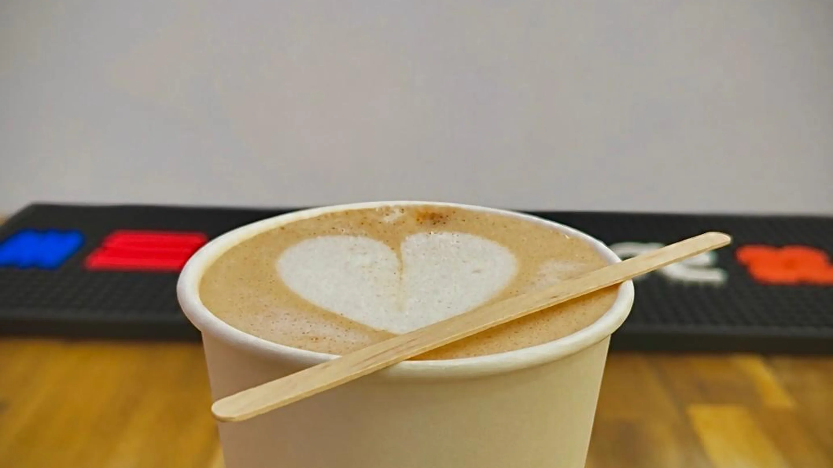 Gros plan d'un gobelet en carton blanc posé sur un comptoir en bois clair. La mousse de lait dessine un cœur à la surface du café, un bâtonnet en bois est posé en travers du bord. Derrière, flous, un tapis de bar noir et l'enseigne néon rose et bleue GROF.
