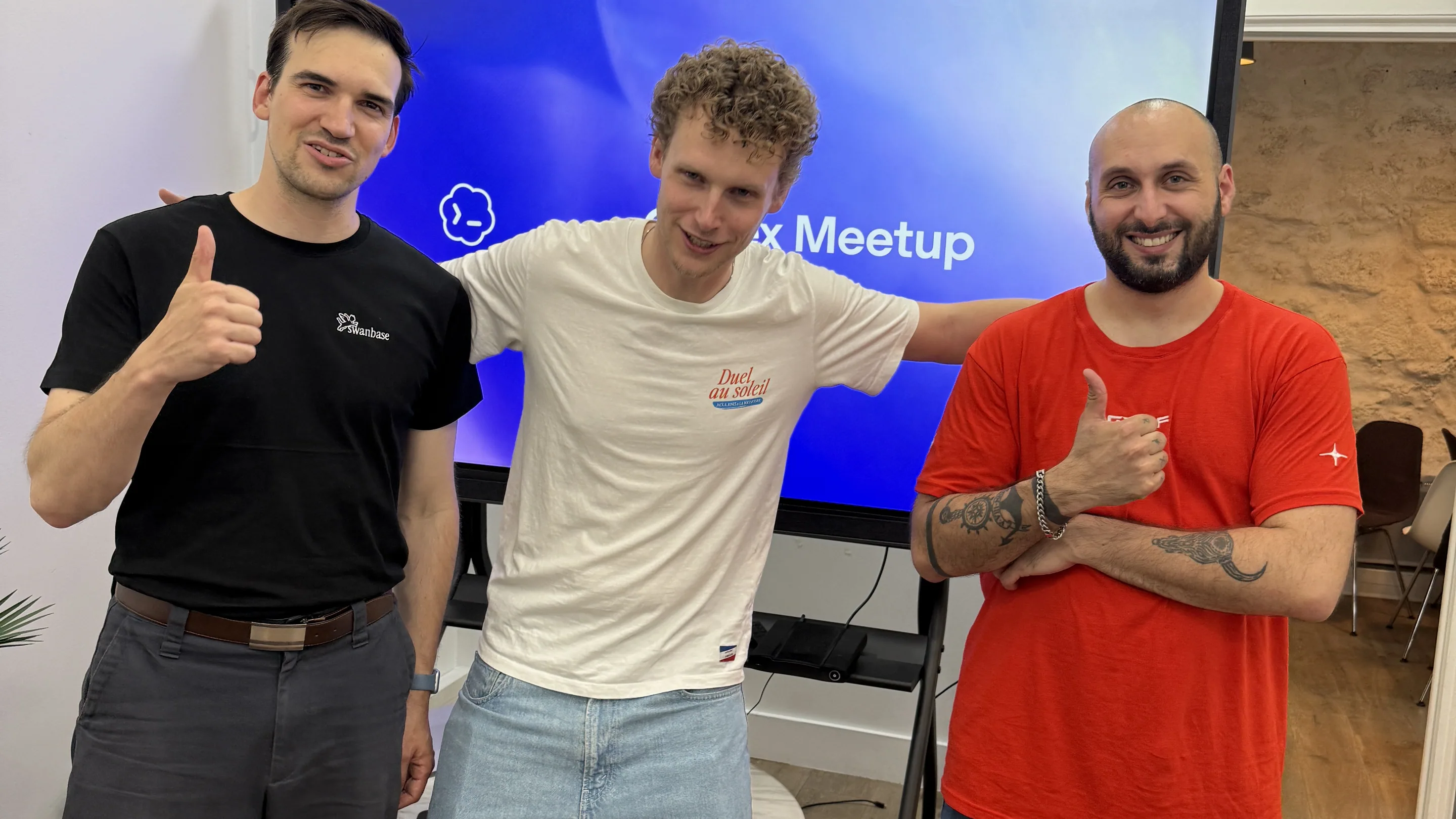 Trois hommes posent côte à côte, pouces levés et sourire aux lèvres, devant un grand écran bleu où se lit le mot « Meetup ». La salle a un parquet clair, un mur de pierre apparente sur la droite et quelques chaises empilées au fond. Celui de gauche porte un t-shirt noir floqué Swanbase, celui du milieu un t-shirt blanc et un short en jean, celui de droite un t-shirt rouge.
