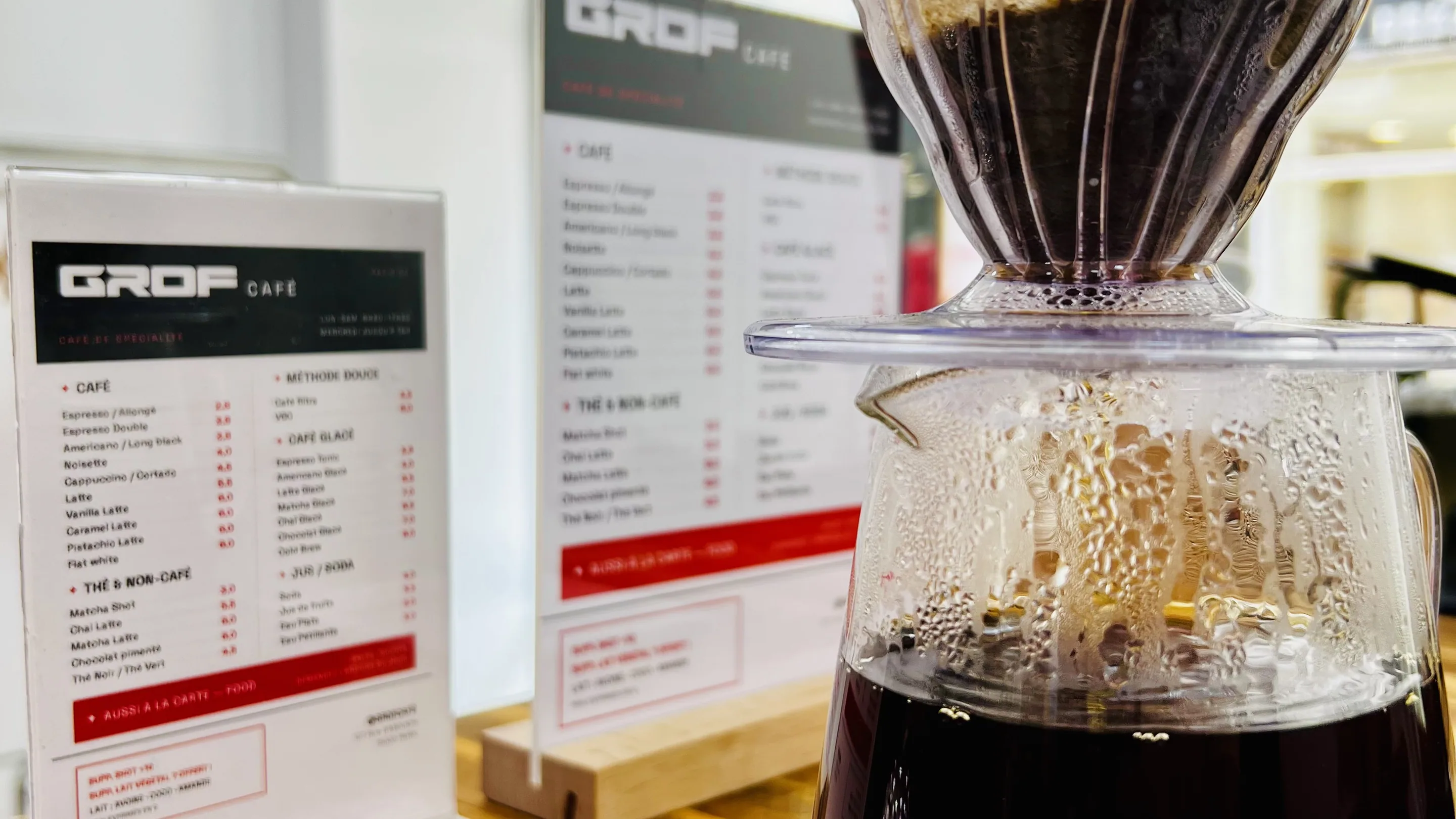 Un café en cours d'infusion sur le comptoir en bois de GROF Café : un dripper conique en verre posé sur une carafe, elle-même sur une balance noire, le marc gonflé sous l'eau chaude et la carafe déjà à moitié pleine. Derrière, deux cartes du menu GROF CAFÉ posées sur leur support, devant la vitrine.