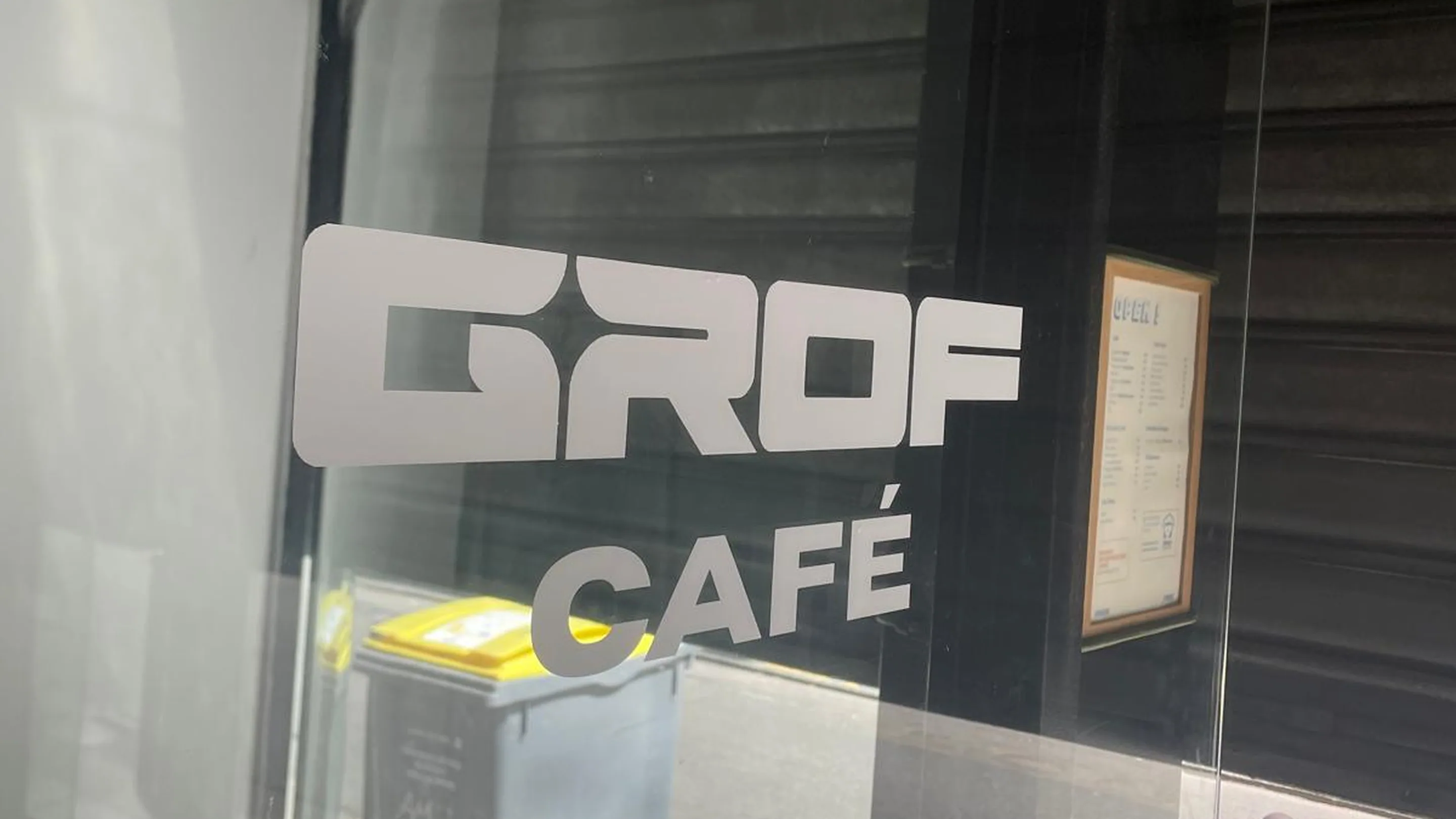 Porte vitrée d'entrée de GROF CAFÉ vue depuis le trottoir, avec le logo GROF CAFÉ en grandes lettres blanches collées sur le verre ; le vitrage reflète la rue et la façade d'en face, et une carte encadrée est affichée à droite de l'entrée.
