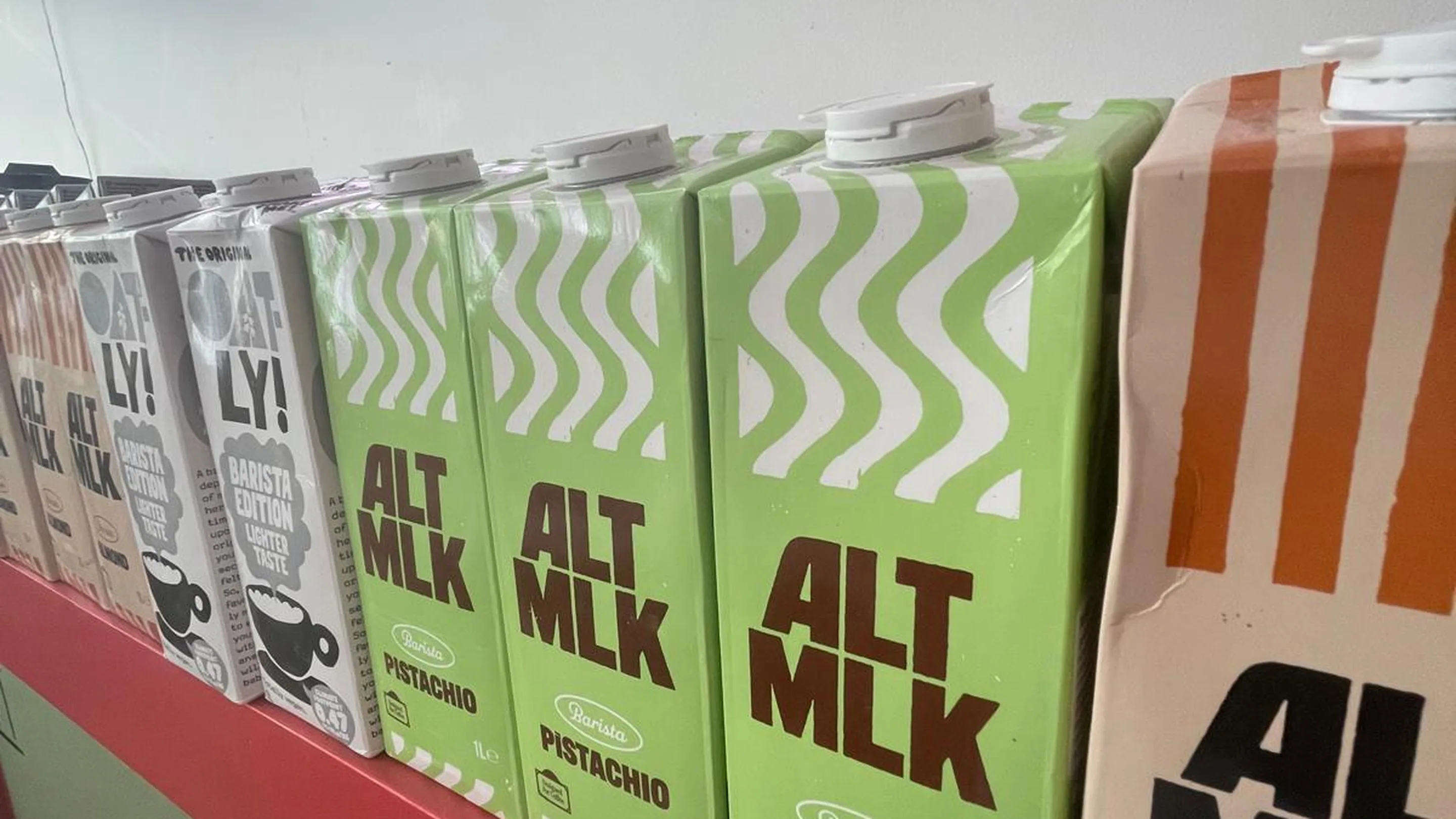 Briques de laits végétaux alignées sur le rebord du comptoir rouge de GROF : deux Oatly Barista à l'avoine, trois Alt Mlk pistache vert pomme et une Alt Mlk amande. Au-dessus, sur le mur blanc, l'enseigne néon rose et bleue GROF.