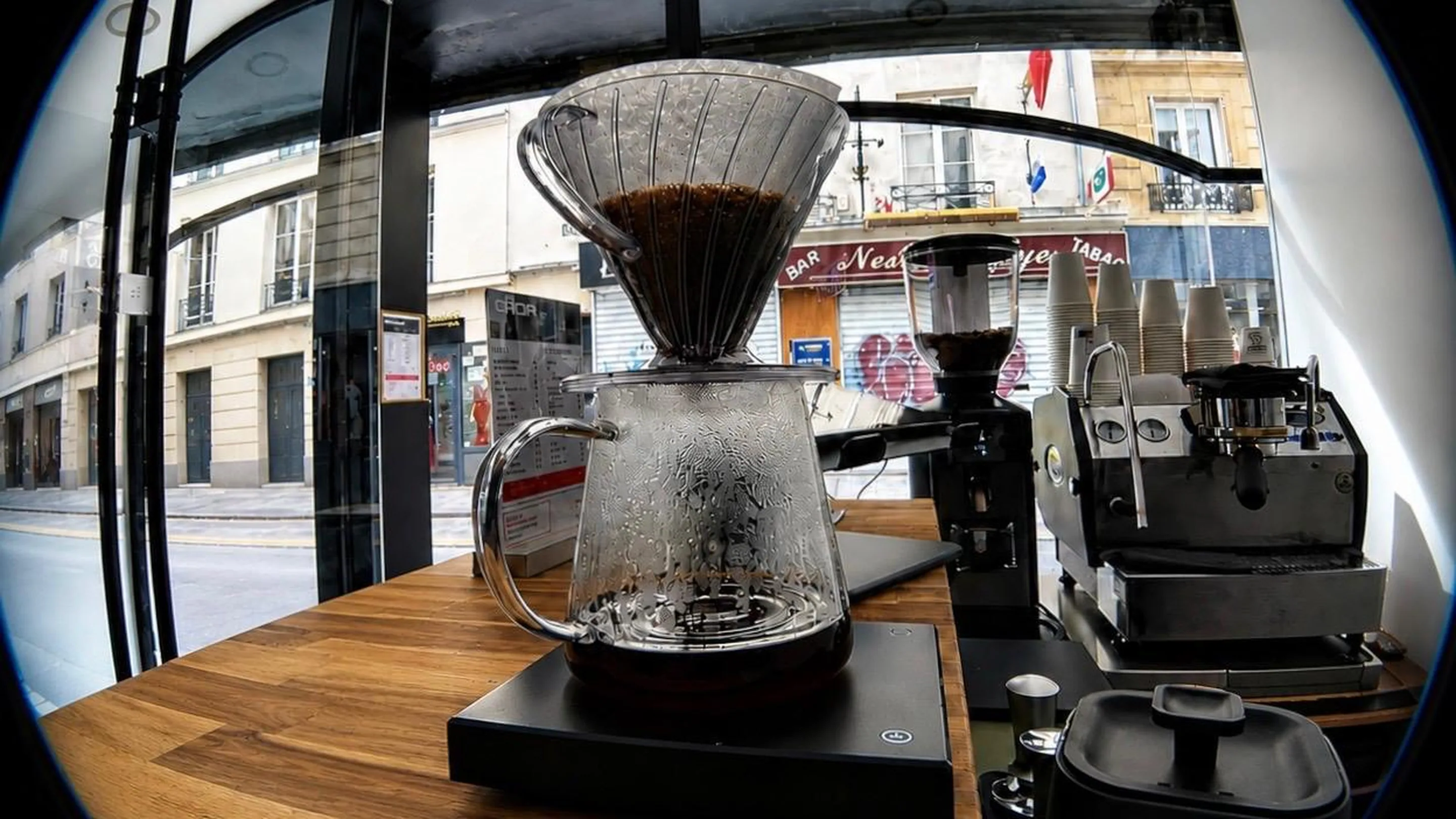 Vue en fisheye du comptoir en bois de GROF : un café filtre en cours d'infusion, dripper en verre posé sur une carafe et une balance noire ; derrière, le moulin, la machine à espresso et une pile de gobelets. Par la grande vitrine, la rue d'Aboukir et les immeubles d'en face.