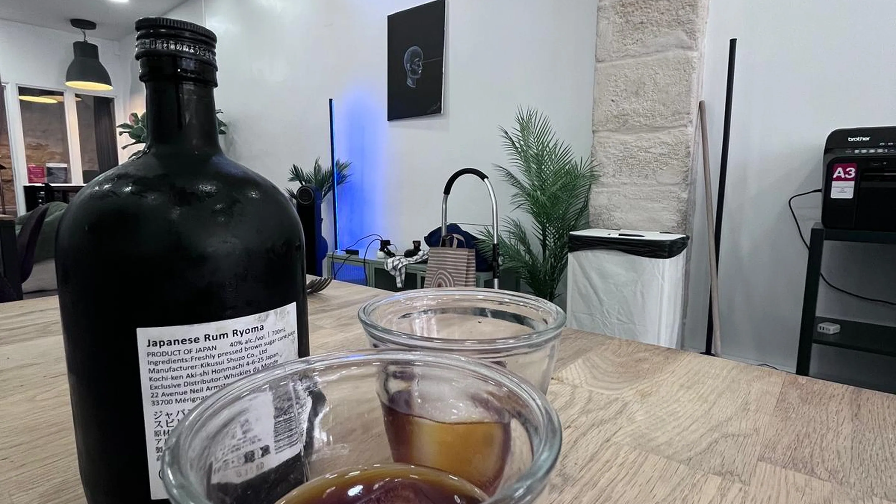 Deux verres transparents posés sur une grande table en bois clair, remplis d'un café glacé très sombre sur un gros glaçon. Derrière, une bouteille noire de rhum japonais Ryoma et la salle du café : mur blanc, pilier en pierre apparente, plante verte, suspensions noires et un petit tableau au mur.