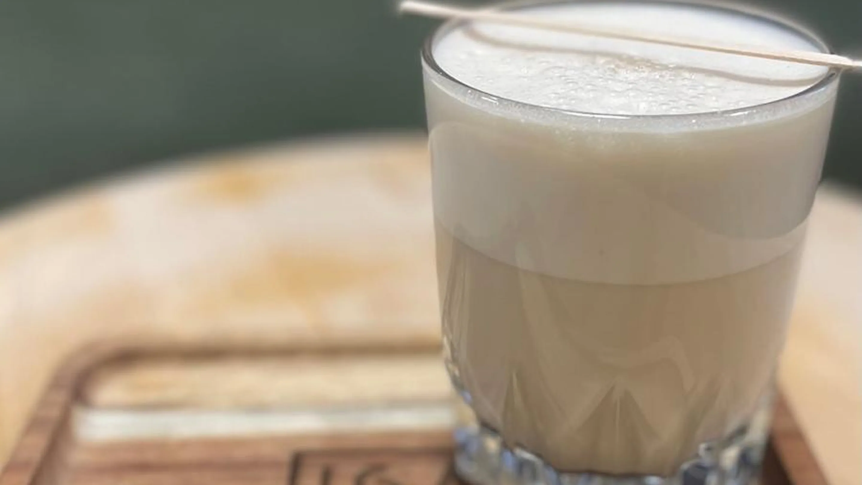 Gros plan d'un latte shot servi dans un petit verre à facettes, posé sur une planche en bois gravée d'un logo. Deux couches nettes dans le verre : café au lait clair en bas, mousse blanche et épaisse au-dessus, une fine touillette en bois posée en travers. Arrière-plan flou aux tons vert et bleu.